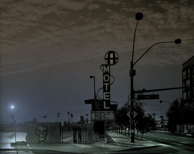 A moody photograph of a motel sign and street signs at an intersection.
