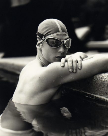 A black and white photograph of a swimmer in cap and goggles leaning on the pool’s edge, water rippling around shoulder.