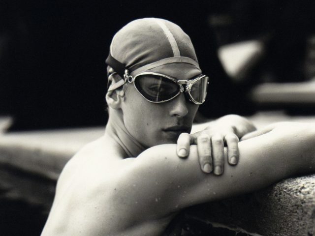 A black and white photograph of a swimmer in cap and goggles leaning on the pool’s edge, water rippling around shoulder.