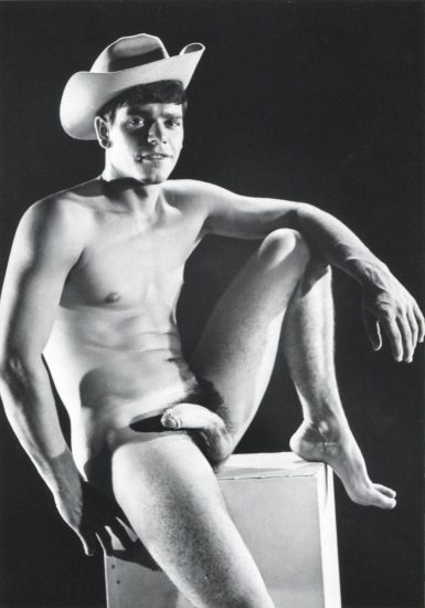 A black and white portrait of a nude young man sitting on a pedestal wearing a cowboy hat.