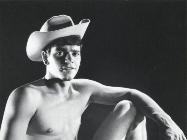 A black and white portrait of a nude young man sitting on a pedestal wearing a cowboy hat.