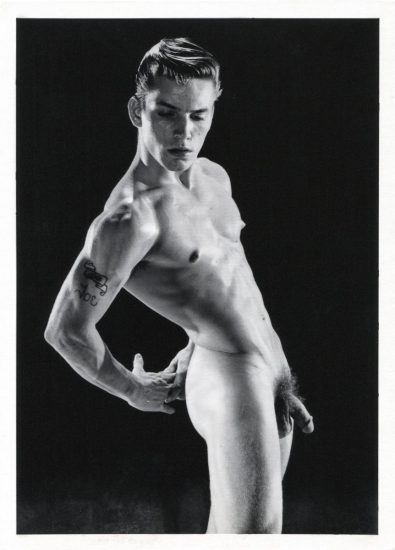 A black and white photograph of a handsome nude young man in a studio setting.