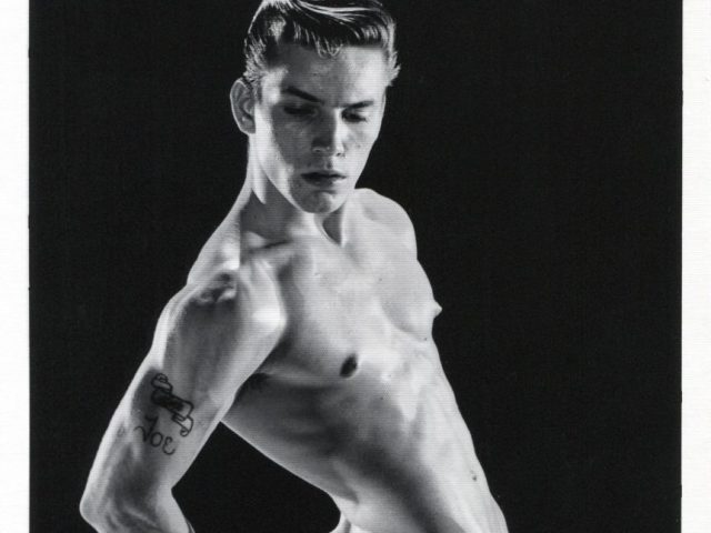 A black and white photograph of a handsome nude young man in a studio setting.