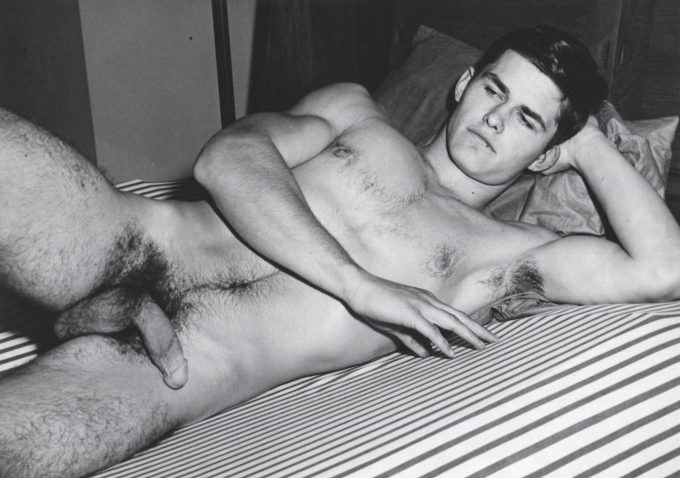 A black and white photograph of a nude young man lying on a striped bed.