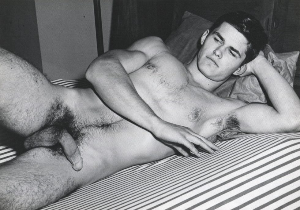 A black and white photograph of a nude young man lying on a striped bed.