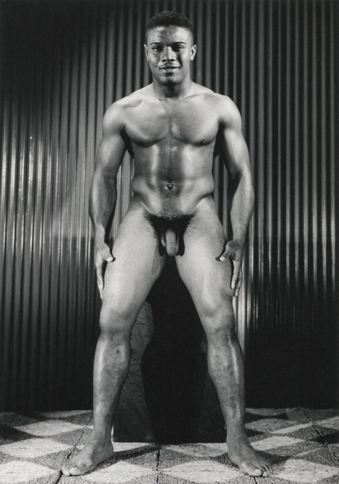 A black and white photograph of a nude young man standing on tiles in front of corrugated metal.