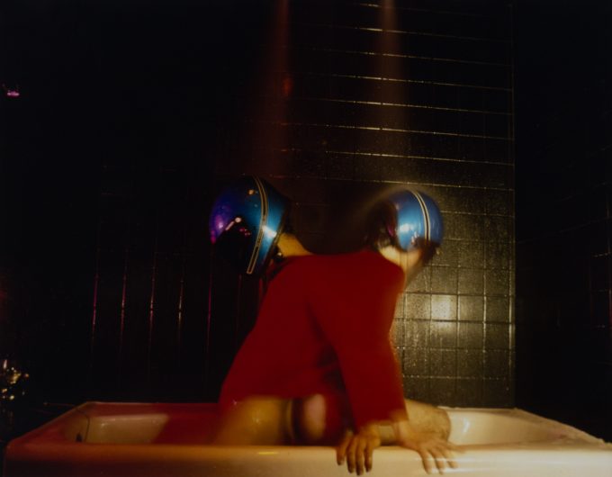 A photograph of two figures in a bathtub wearing motorcycle helmets.