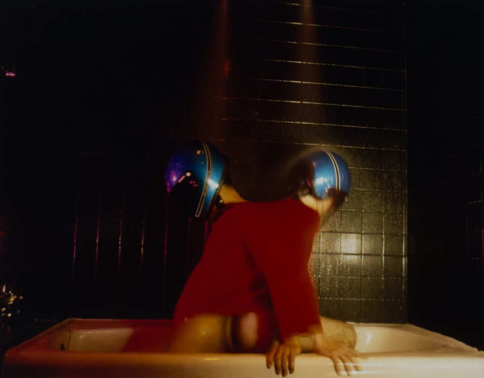 A photograph of two figures in a bathtub wearing motorcycle helmets.