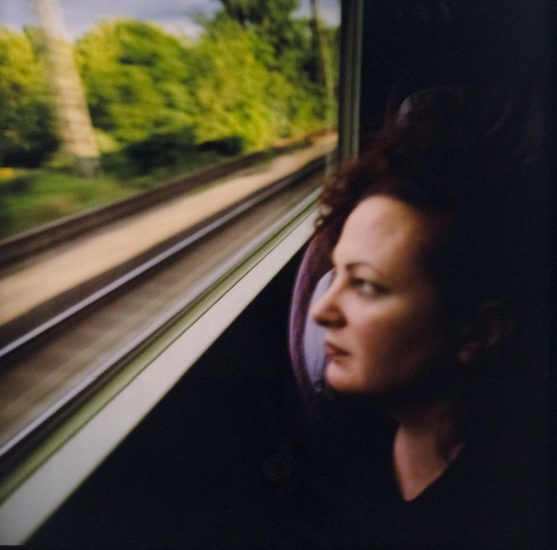 A person with curly red hair gazes out a train window as blurred tracks and green trees rush past.
