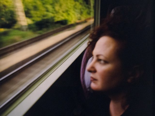 A person with curly red hair gazes out a train window as blurred tracks and green trees rush past.
