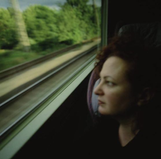 A person with curly red hair gazes out a train window as blurred tracks and green trees rush past.