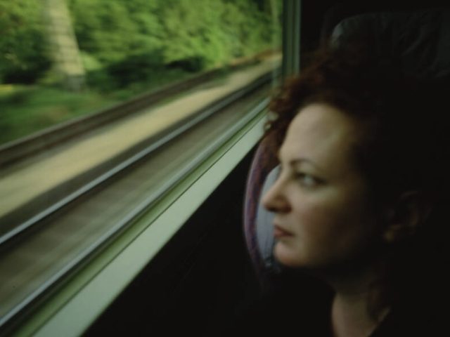 A person with curly red hair gazes out a train window as blurred tracks and green trees rush past.
