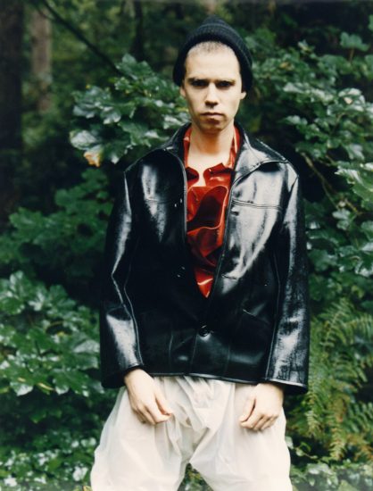 A photograph of a man in a black beanie, red shirt, black leather jacket, and transparent plastic pants.