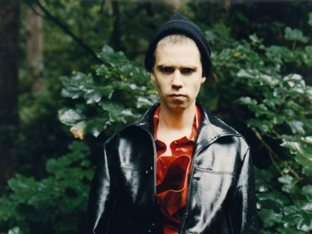 A photograph of a man in a black beanie, red shirt, black leather jacket, and transparent plastic pants.