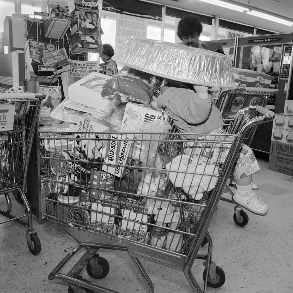 A black and white photograph of a child in a shopping cart in a supermarket.