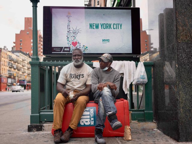 A colorful photograph of two older men sitting in front of a subway entrance.