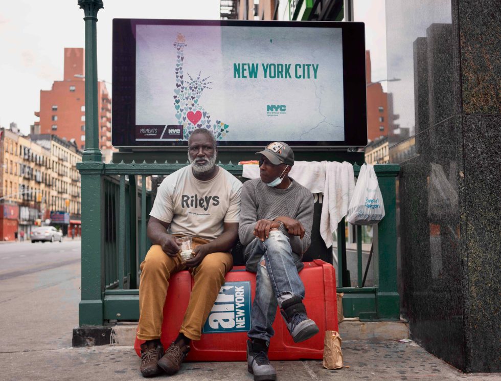 A colorful photograph of two older men sitting in front of a subway entrance.
