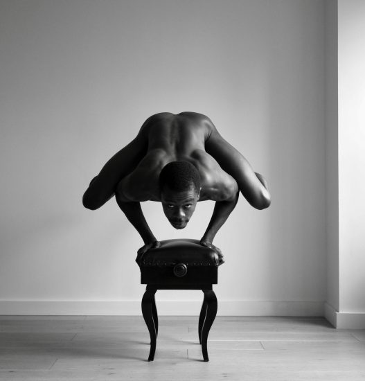 A black and white photograph of a nude man balancing on his hands on top of a piano bench.