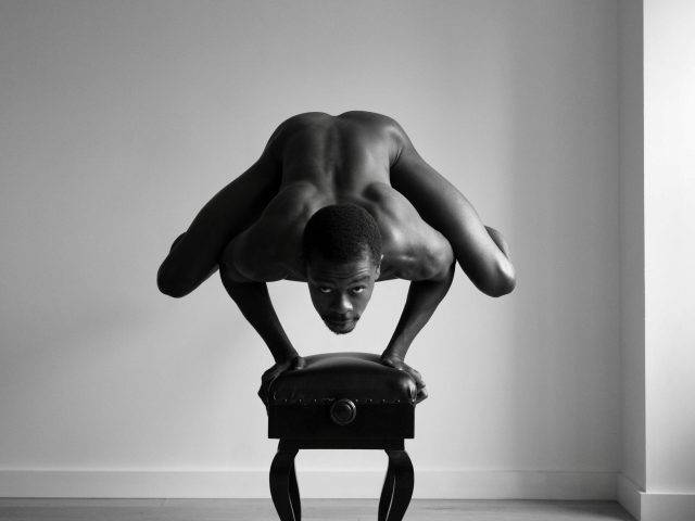 A black and white photograph of a nude man balancing on his hands on top of a piano bench.