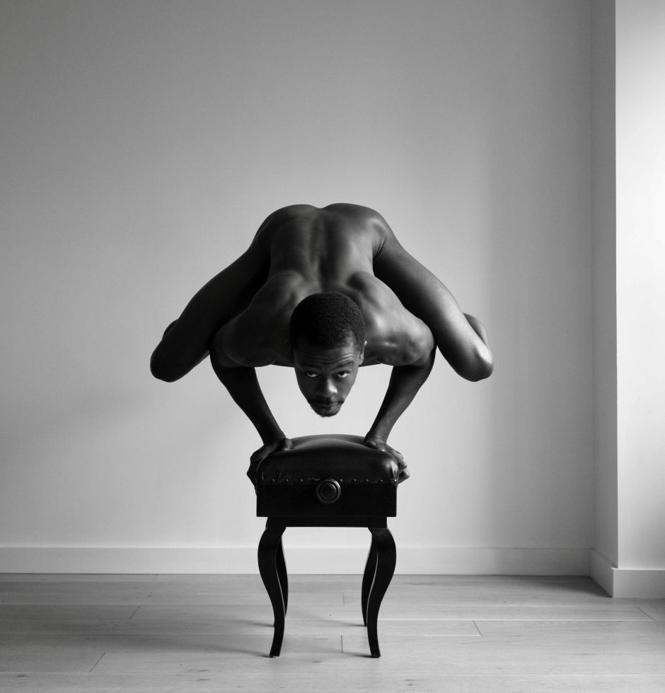 A black and white photograph of a nude man balancing on his hands on top of a piano bench.