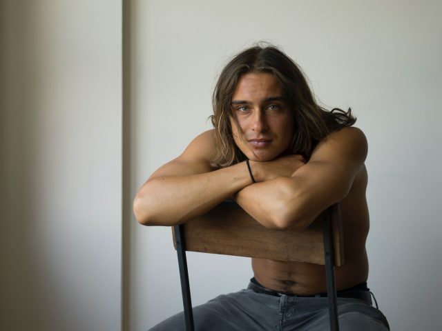 A photograph of a handsome man with long hair sitting in a chair.
