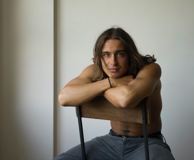 A photograph of a handsome man with long hair sitting in a chair.