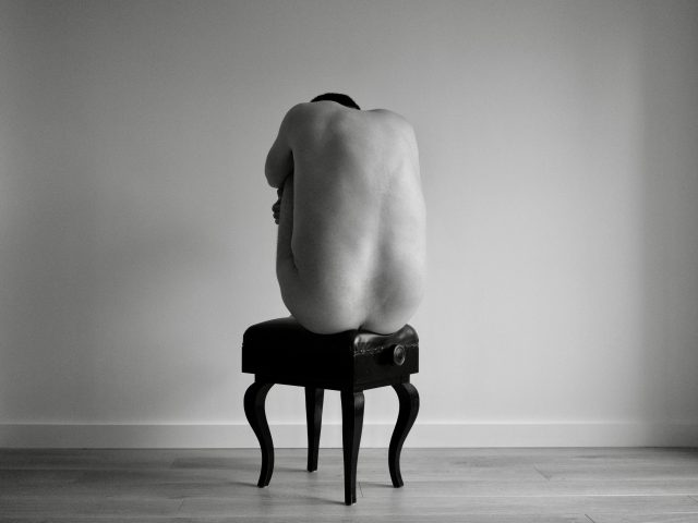 A black and white photograph of a nude man sitting on a piano bench.