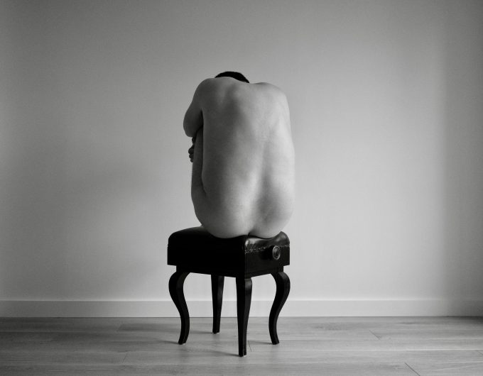A black and white photograph of a nude man sitting on a piano bench.