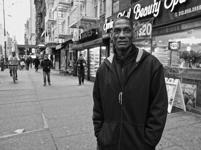 A black and white photograph of a man standing outside a storefront on a city street.