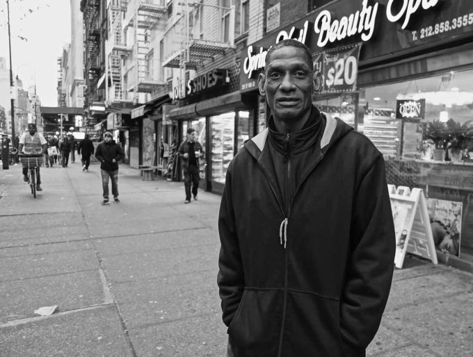 A black and white photograph of a man standing outside a storefront on a city street.