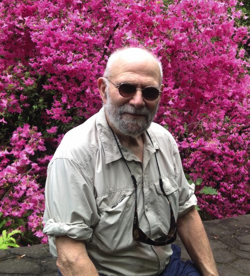 A color photograph of an older man smiling in sunglasses in front of pink blooming flowers.