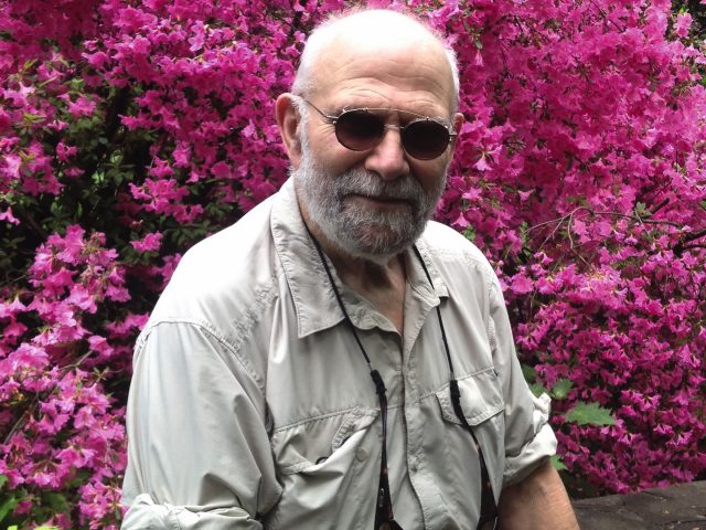 A color photograph of an older man smiling in sunglasses in front of pink blooming flowers.