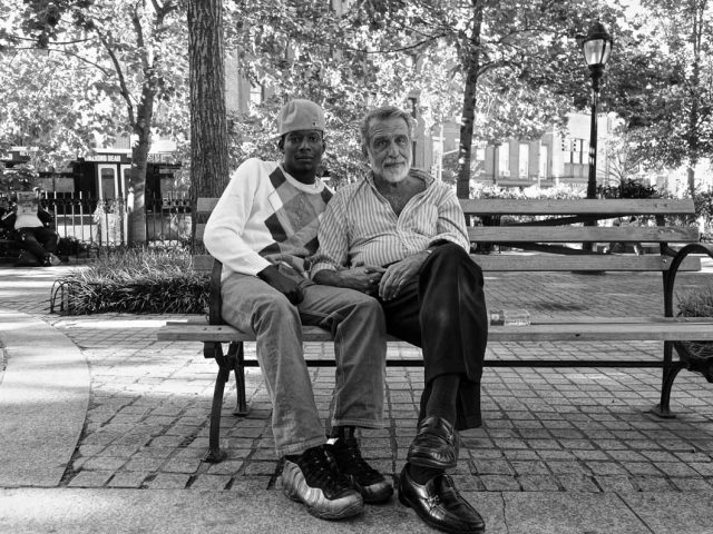 A black and white photograph of two men sitting on a bench in a park.