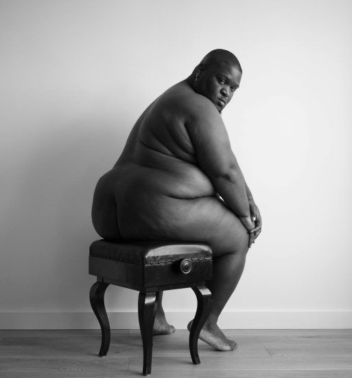 A black and white photograph of a chubby nude man sitting on a piano bench.