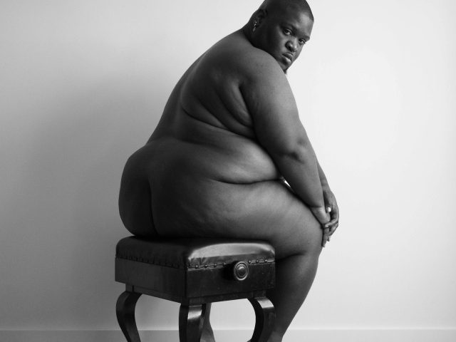 A black and white photograph of a chubby nude man sitting on a piano bench.