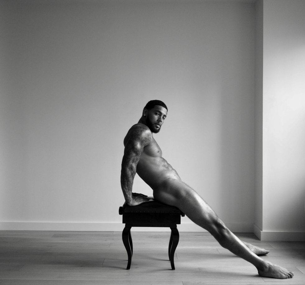 A black and white portrait of a nude man sitting on a piano bench.