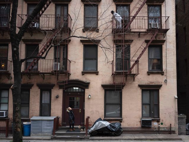 A photograph of a person standing at the front door of an apartment building calling up to someone on the third floor.