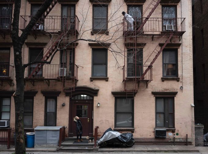 A photograph of a person standing at the front door of an apartment building calling up to someone on the third floor.