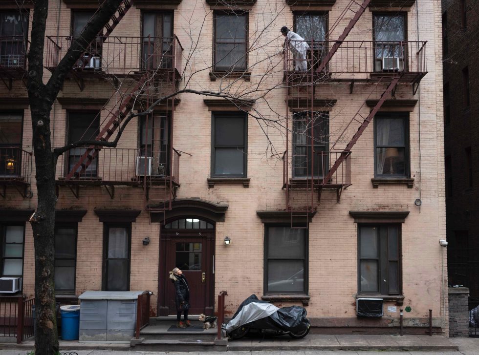 A photograph of a person standing at the front door of an apartment building calling up to someone on the third floor.