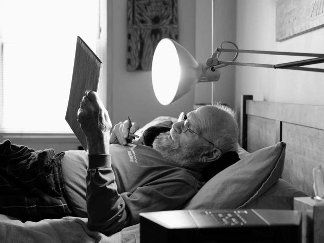 A black and white image of an older man lying in bed reading.