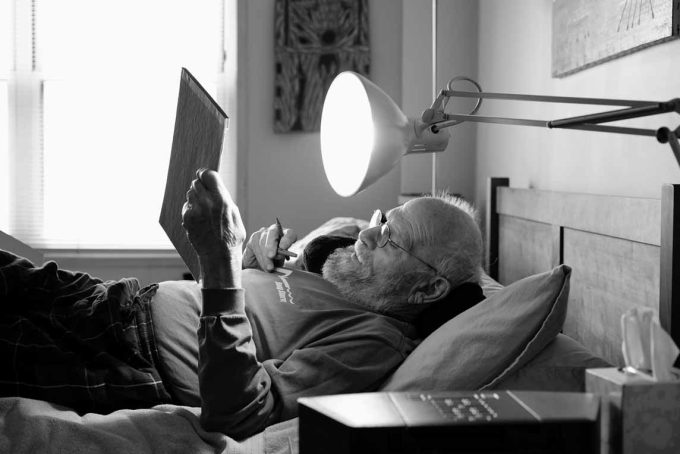A black and white image of an older man lying in bed reading.