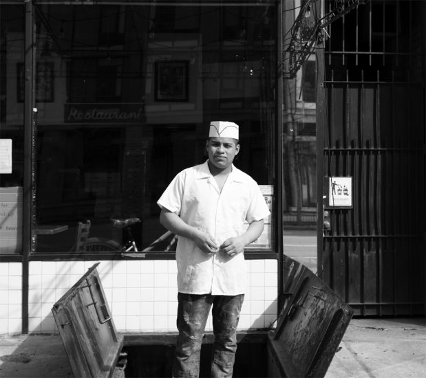 A black and white photograph of a line cook standing in front of a storefront.