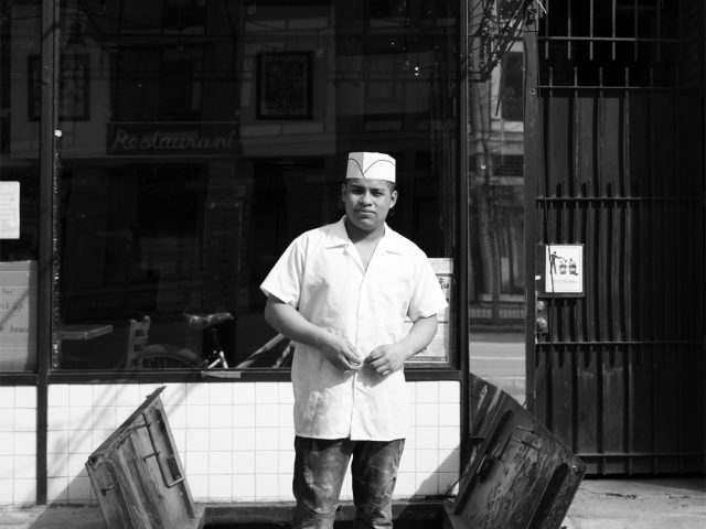 A black and white photograph of a line cook standing in front of a storefront.