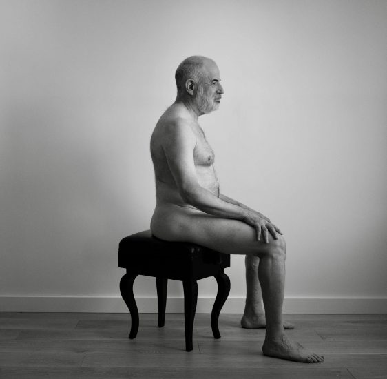 A black and white photograph of a nude man on a piano bench.