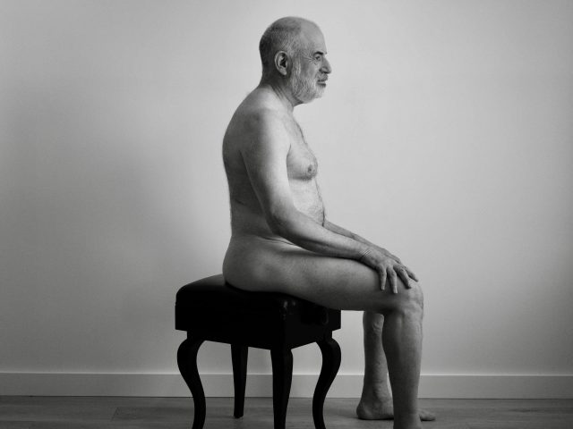 A black and white photograph of a nude man on a piano bench.