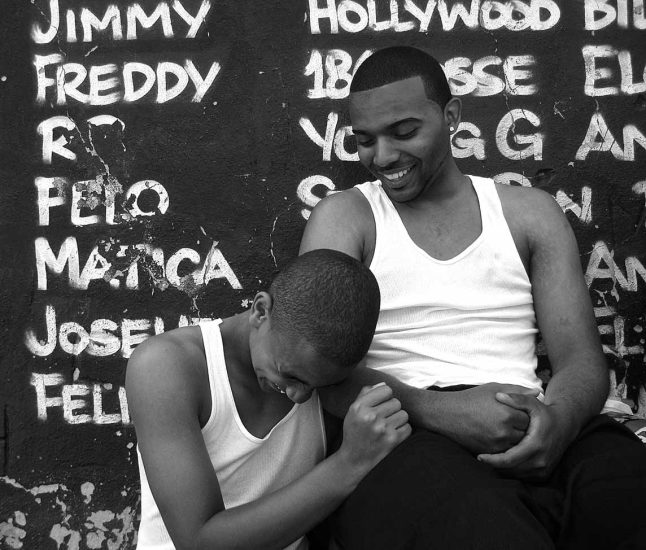A black and white photograph of two men sitting in front of a grafitti-covered wall.