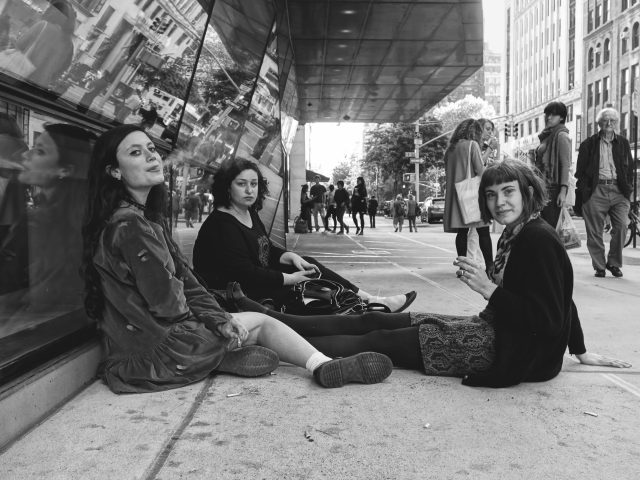A black and white photograph of three young women sitting on the sidewalk smoking cigarettes.