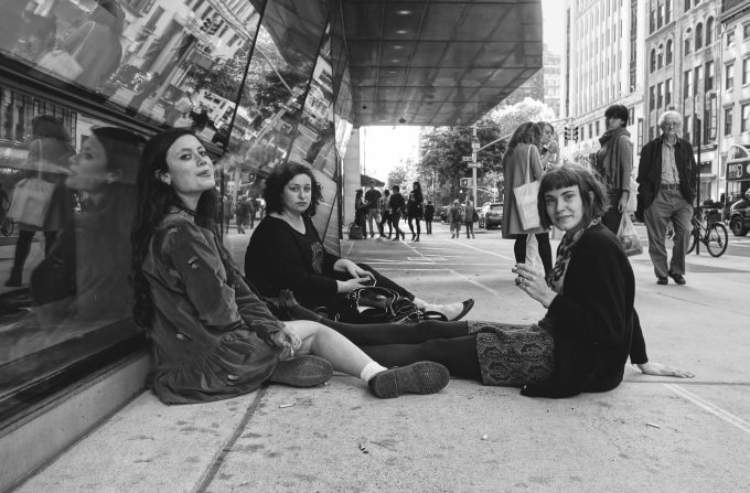 A black and white photograph of three young women sitting on the sidewalk smoking cigarettes.