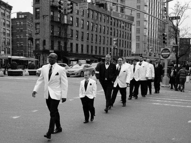 A group of men, young and old, walk across a crosswalk in white and black suits.