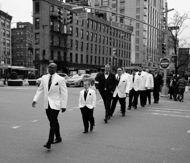 A group of men, young and old, walk across a crosswalk in white and black suits.